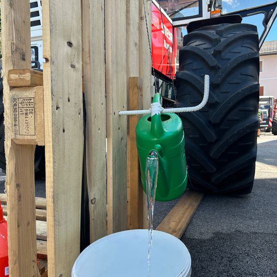 Ein grüner Gießkanister ist an einem Holzzaun befestigt, wobei Wasser in einen weißen Eimer fließt. Ein Traktor mit einem großen Reifen steht hinter dem Zaun.