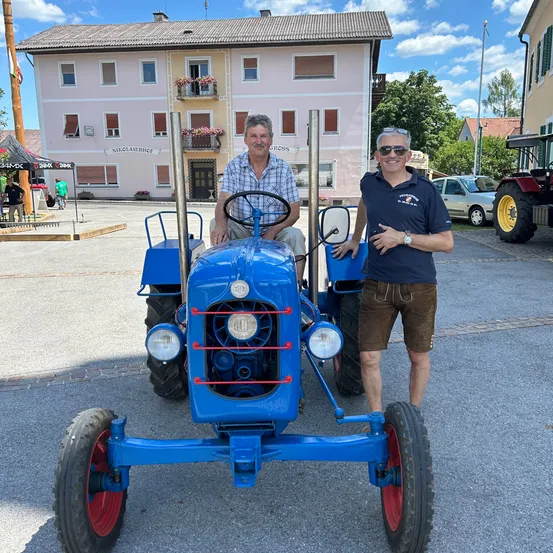 Zwei Männer stehen neben einem blauen Traktor vor einem Gebäude mit Blumen auf dem Balkon. Dahinter sind Autos geparkt.