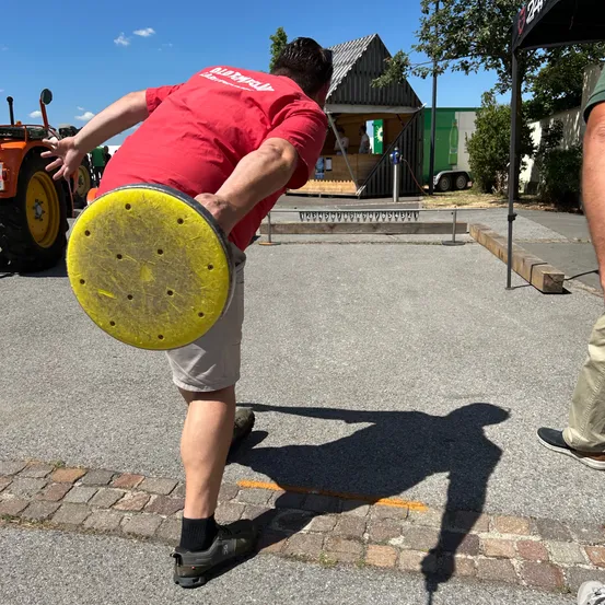 Ein Mann in einem roten T-Shirt hält einen gelben Scheiben im Freien und bereitet sich darauf vor, sie zu werfen. Er steht in der Nähe eines Traktors und einer kleinen Hütte.