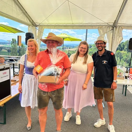 Vier Personen stehen in einem Zelt unter einem klaren Himmel. Ein Mann in einem roten Shirt hält eine Geschenkbox. Zwei Frauen und ein weiterer Mann stehen neben ihm und lächeln. Dahinter steht ein Tisch mit einer Aufbewahrungsbox.