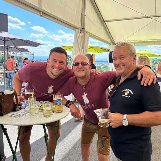 Drei Männer lächeln und posieren für ein Foto unter einem Zelt mit Sonnenschirmen. Ein Mann hält ein Glas Bier in der Hand.