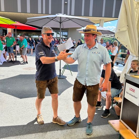 Zwei Männer in traditioneller bayerischer Kleidung tauschen einen Händedruck im Freien aus. Hinter ihnen versammeln sich Leute unter Schirmen, einige mit Getränken in der Hand. Ein Mann trägt einen Strohhut und hält ein Mikrofon. Ein Stand mit der Aufschrift 'LOSE' ist in der Nähe, was auf eine mögliche Veranstaltung oder einen Wettbewerb hindeutet.