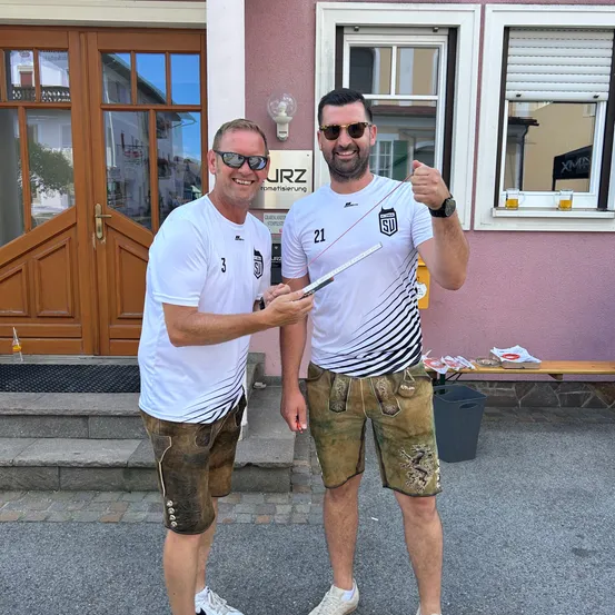 Zwei Männer in Sportbekleidung, lächelnd und posieren für ein Foto vor einem Gebäude. Ein Mann hält einen Stock. Dahinter befinden sich eine Bank, ein Mülleimer und ein Schild.