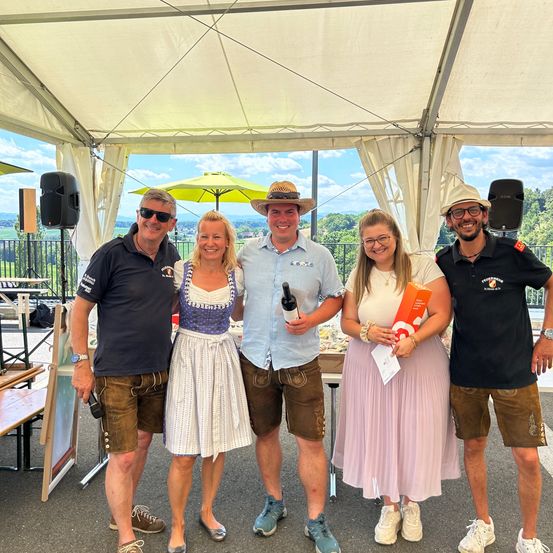 Fünf Menschen stehen lächelnd unter einem Zelt. Ein Mann hält eine Flasche. Hinter ihnen steht ein Tisch mit einem Lautsprecher. Im Hintergrund sind Bäume und ein Zaun zu sehen.