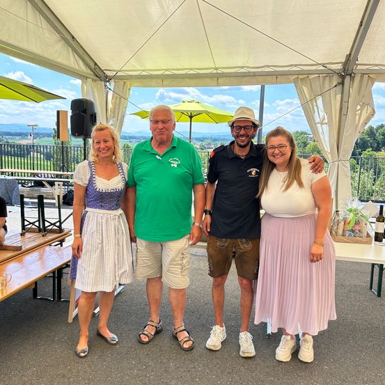Vier Personen stehen unter einem Zelt mit einer malerischen Aussicht im Hintergrund. Eine Frau in einem traditionellen Kleid und ein Mann in einem grünen Hemd stehen links, während ein Mann und eine Frau in lässiger Kleidung rechts stehen.