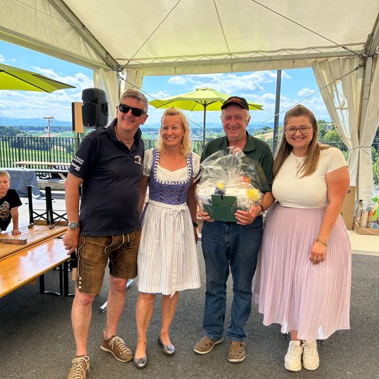 Vier Menschen stehen unter einem Zelt mit einer landschaftlichen Aussicht hinter ihnen. Zwei Männer und zwei Frauen, alle lächeln und posieren für ein Foto. Der Mann auf der linken Seite trägt eine Uhr und Ledershorts, während die Frau in der Mitte einen Geschenkkorb hält.