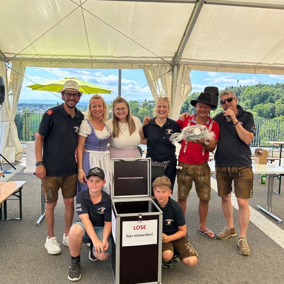 Eine Gruppe von Menschen steht in einem Zelt zusammen und lächelt für ein Foto. Ein Mann hält einen Blumenstrauß und spricht in ein Mikrofon. Eine schwarze Kiste mit der Aufschrift LOSE steht in der Mitte.