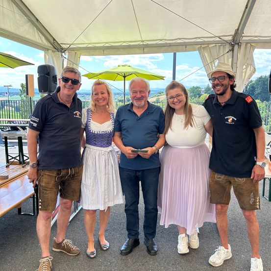 Fünf Menschen stehen unter einem Zelt, lächeln und posieren für ein Foto. Die Frau in der Mitte trägt ein traditionelles Kleid, und zwei Männer tragen Uniformen. Sie befinden sich in einem Außenbereich mit einer Holzbank und Sonnenschirmen.