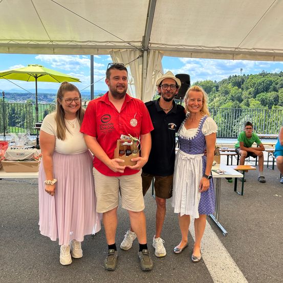 Vier Personen stehen unter einem weißen Zelt. Zwei Frauen und zwei Männer. Der Mann in der Mitte hält eine Box. Hinter ihnen sind Tische und eine Bank. Bäume und Berge sind im Hintergrund.