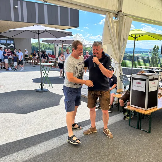 Zwei Männer stehen und reden, während sie sich an den Händen halten. Einer hält ein Mikrofon. In der Nähe steht eine Box mit der Aufschrift 'LOSE'. Im Hintergrund sind mehrere Menschen versammelt, einige sitzen und andere stehen. Es sind Sonnenschirme und Tische aufgestellt.