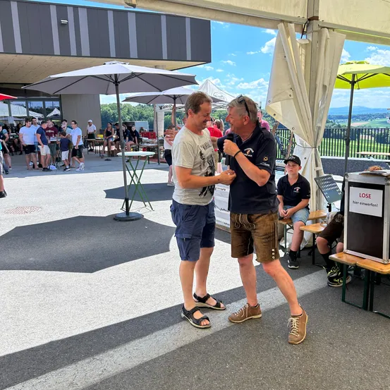 Zwei Männer unterhalten sich in der Nähe eines Regenschirms und eines Podiums in einem großen Veranstaltungsraum. Menschen versammeln sich unter Regenschirmen, und ein Schild steht LOSE.