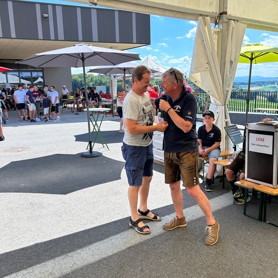 Zwei Männer unterhalten sich in der Nähe eines Regenschirms und eines Podiums in einem großen Veranstaltungsraum. Menschen versammeln sich unter Regenschirmen, und ein Schild steht LOSE.