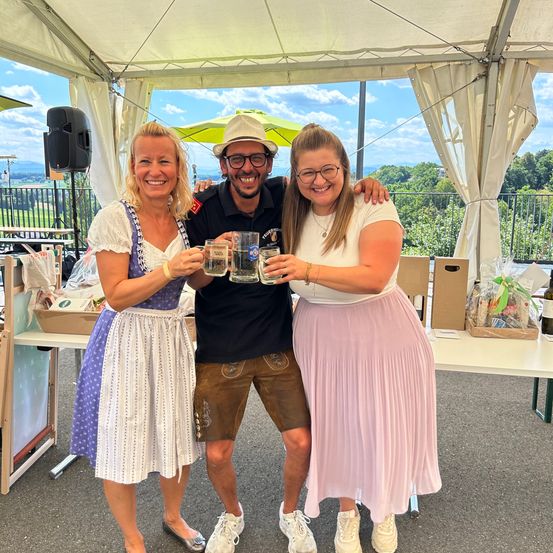 Drei Personen stehen unter einem Zelt. Eine Frau links trägt ein traditionelles Kleid, ein Mann in der Mitte trägt eine Brille und einen Hut und eine Frau rechts trägt einen rosa Rock. Sie halten alle ein Glas Bier.