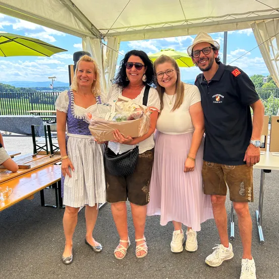 Vier Menschen stehen unter einem Zelt und lächeln für ein Foto. Eine Frau hält einen Geschenkkorb. Hinter ihnen sind Picknicktische aufgebaut. Bäume und Berge sind im Hintergrund sichtbar.