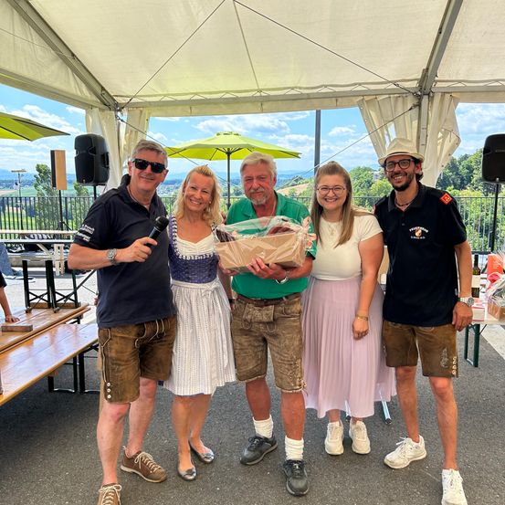 Vier Menschen stehen unter einem Zelt und lächeln. Der Mann auf der linken Seite hält ein Mikrofon und spricht. Der Mann in der Mitte hält einen Geschenkkorb. Die Frau in der Mitte trägt ein traditionelles Kleid. Hinter ihnen stehen Bänke, Lautsprecher und Tische.