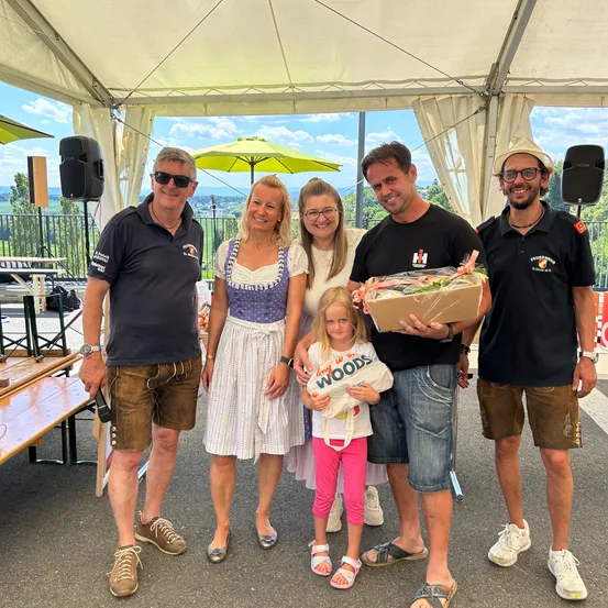 Eine Familie mit einem kleinen Mädchen steht unter einem Zelt und posiert für ein Foto. Der Mann auf der linken Seite trägt eine Sonnenbrille und eine Halskette. Der Mann auf der rechten Seite hält eine Box, und ein Mädchen hält eine Tasche. Hinter ihnen befinden sich Lautsprecher, ein Tisch und ein Zaun.