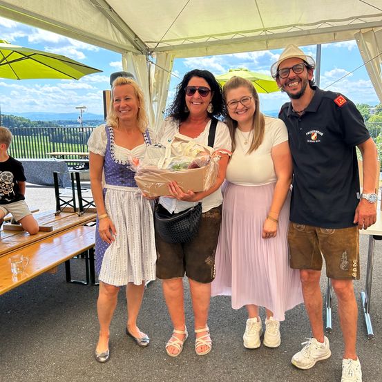 Vier Personen stehen unter einem Zelt, mit einem Jungen, der auf einer Bank hinter ihnen sitzt. Die drei Frauen tragen Brillen und halten einen Korb, während der Mann lächelt.