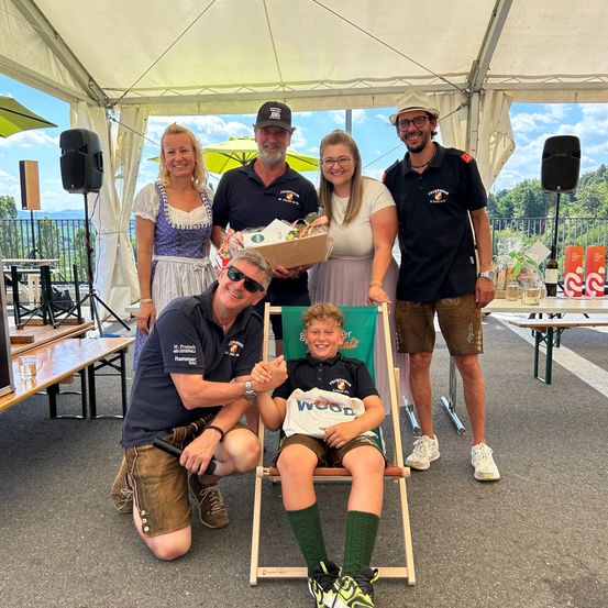 Eine Gruppe von Erwachsenen und einem Kind stehen und hocken unter einem Zelt und lächeln für ein Foto. Die Erwachsenen sind in Freizeitkleidung gekleidet. Das Kind hält eine Tasche mit dem Wort Wood.