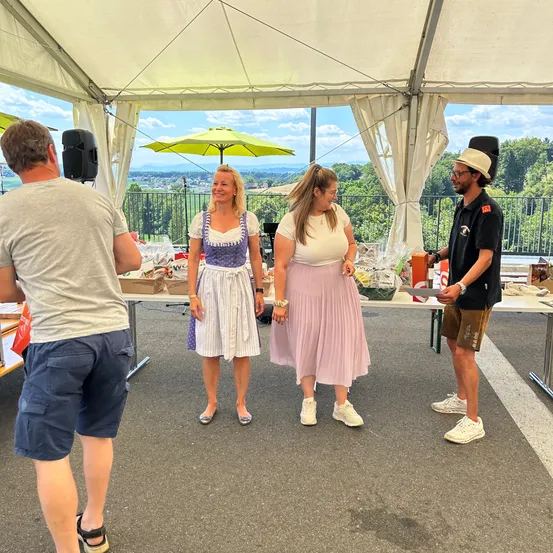 Unter einem Zelt bereiten sich zwei Frauen und ein Mann auf eine Veranstaltung vor. Die Frauen tragen Kleider und der Mann hält ein Geschenk. Dahinter steht ein Tisch mit Gegenständen. Draußen gibt es eine Terrasse und Bäume.