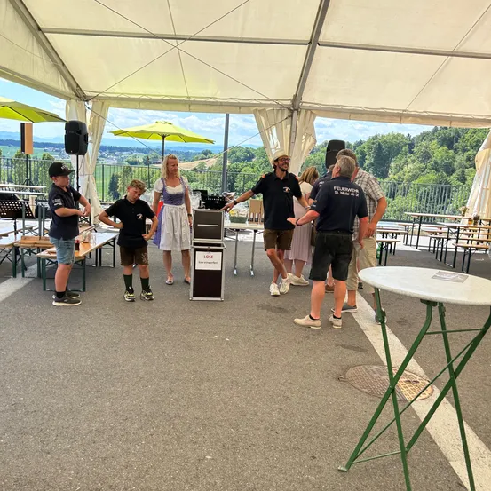 Eine Gruppe von Menschen steht unter einem Zelt mit Tischen und Stühlen. Ein Mann zeigt auf eine Box mit der Aufschrift VERLORE. Im Hintergrund sind Bäume und Berge zu sehen.