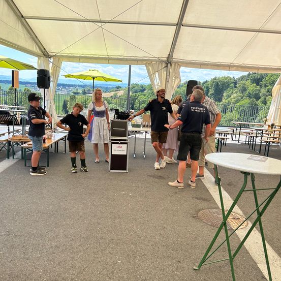 Eine Gruppe von Menschen steht unter einem Zelt mit Tischen und Stühlen. Ein Mann zeigt auf eine Box mit der Aufschrift VERLORE. Im Hintergrund sind Bäume und Berge zu sehen.