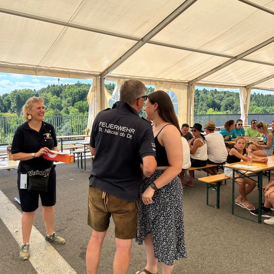 Eine Gruppe von Menschen genießt eine Mahlzeit an einem Tisch im Freien unter einem Zelt. Eine Frau hält eine Box, während ein Mann und eine andere Frau in der Nähe sprechen. Bäume und ein Zaun sind im Hintergrund zu sehen.