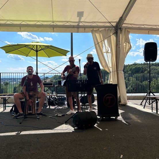 Drei Männer spielen auf einer Freiluftbühne unter einem Zelt. Einer spielt Gitarre, ein anderer singt und der dritte hält ein Mikrofon. Ein gelber Regenschirm bietet Schatten.