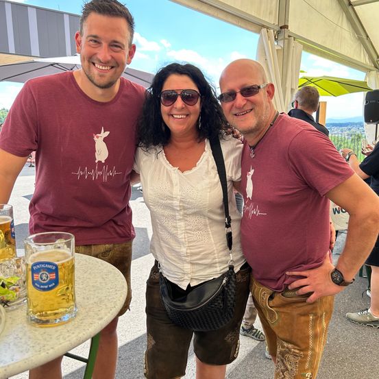 Drei Personen lächeln für ein Foto bei einer Outdoor-Veranstaltung. Eine Frau steht zwischen zwei Männern, alle in Freizeitkleidung gekleidet. Hinter ihnen befinden sich Zelte, ein Tisch mit einem Bierkrug und herumstehende Personen.