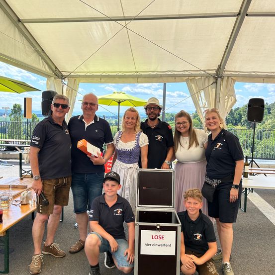 Eine Gruppe von Menschen steht unter einem Zelt mit einem Schild, auf dem LOSE steht. Sie posieren für ein Foto. Dahinter befinden sich Tische, Bänke und Bäume.