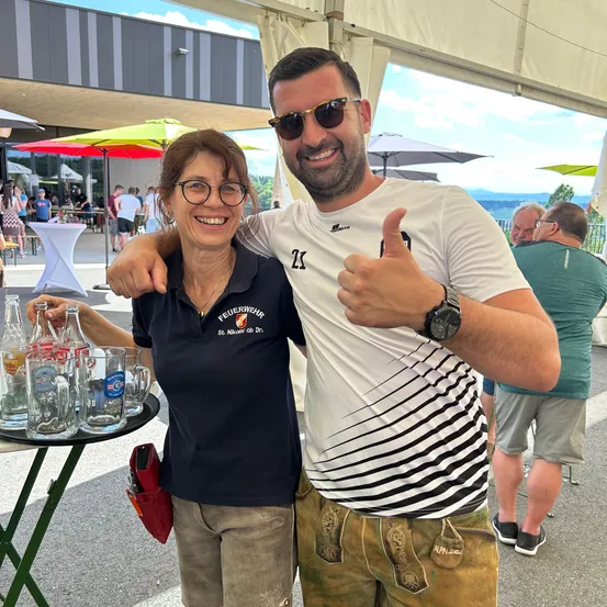 Ein Mann und eine Frau lächeln für ein Foto bei einer Veranstaltung im Freien, mit Biergläsern und einem Tisch dahinter. Dahinter sitzen und stehen Personen bei Tischen und Schirmen.