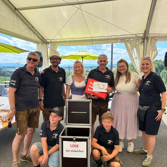 Eine Gruppe von Menschen versammelt sich unter einem Zelt, lächelt und posiert für ein Foto. Sie halten eine rote Box mit dem Wort Lose. Die Gruppe besteht aus vier Erwachsenen und zwei Kindern. Die Kinder knien vor der Box. Die Frau in der Mitte hält einen Papierbeutel. Das Gebiet ist von weißen Vorhängen umgeben. In der Ferne gibt es Bäume und einen Zaun.