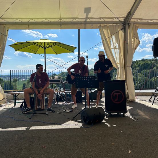 Drei Männer treten auf einer Bühne unter einem Zelt auf. Ein Mann spielt Gitarre, ein anderer sitzt auf einem Schlagzeug und der dritte steht und singt. Hinter ihnen befindet sich ein gelber Schirm und ein schwarzer Lautsprecher.