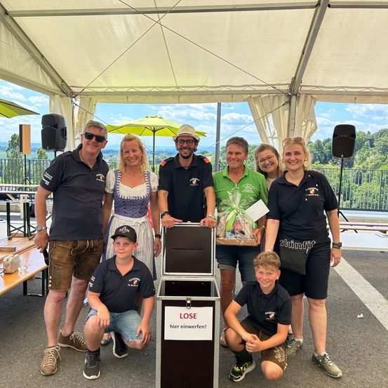 Eine Gruppe von Menschen steht unter einem Zelt mit einer Tombola-Kiste und lächelt. Sie sind in Freizeitkleidung gekleidet. Ein Mann in einem grünen Shirt hält einen Geschenkkorb. Dahinter befindet sich eine Terrasse mit Geländer und ein Lautsprecher auf einem Ständer.