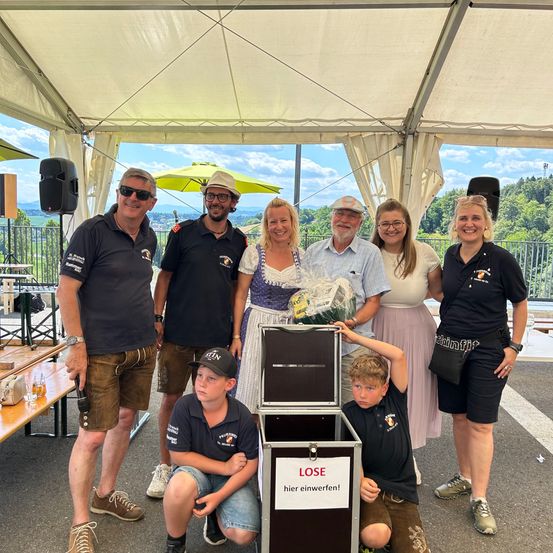Eine Gruppe von Menschen posiert für ein Foto unter einem Zelt, einige mit Hüten, halten eine Box mit der Aufschrift 'LOSE hier einwerfen!'. Dahinter steht eine Bank, ein Lautsprecher und ein Zaun. Der Himmel ist bewölkt.
