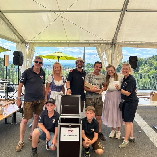 Eine Gruppe von Menschen steht in einem Zelt mit einer großen Kiste vor ihnen, posiert für ein Foto. Sie sind lässig gekleidet und scheinen zu feiern. Hinter ihnen sind ein Lautsprecher und ein gelber Schirm zu sehen.