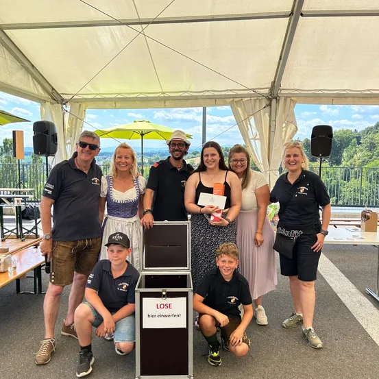 Eine Gruppe von Menschen posiert für ein Foto in einem Zelt. Einige halten Urkunden, und eine Box hat 'LOSE hier einwerfen!' darauf. Eine Frau lächelt und hält eine Urkunde. Ein Tisch mit einer Flasche ist in der Nähe.