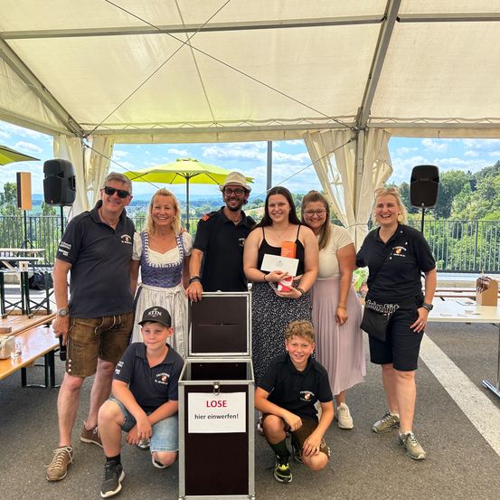 Eine Gruppe von Menschen posiert für ein Foto in einem Zelt. Einige halten Urkunden, und eine Box hat 'LOSE hier einwerfen!' darauf. Eine Frau lächelt und hält eine Urkunde. Ein Tisch mit einer Flasche ist in der Nähe.