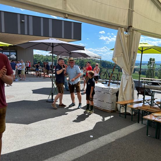 Mehrere Erwachsene stehen unter Schirmen bei einer Outdoor-Veranstaltung. Einige tragen Hüte und Schuhe. Dahinter versammeln sich weitere Menschen, und Fahrräder sind geparkt. Ein Tisch mit weißer Abdeckung steht in der Nähe. Bäume und ein Gebäude sind im Hintergrund.