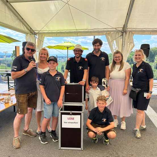 Eine Gruppe von Menschen steht unter einem Zelt. Eine Person hält ein Mikrofon. Ein Junge hockt in der Nähe einer Aufbewahrungsbox. Auf der Box steht das Wort LOSE.