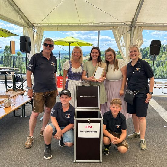 Eine Gruppe von Menschen posiert für ein Foto unter einem Zelt. Zwei Frauen halten ein Buch und lächeln. Zwei Jungen knien neben einer Kiste mit dem Wort 'VERLIEREN' darauf. Ein Mann steht hinter ihnen.