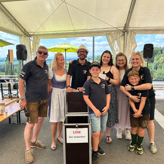 Eine Gruppe von Menschen posiert für ein Foto unter einem Zelt, einer hält eine Spendenbox mit der Aufschrift 'LOSE hier einwerfen!' (werfen Sie Ihr Kleingeld hier rein). Sie tragen lässige Sommerkleidung. Hinter ihnen steht ein Holztisch mit einem Glas, und zwei Lautsprecher sind sichtbar.