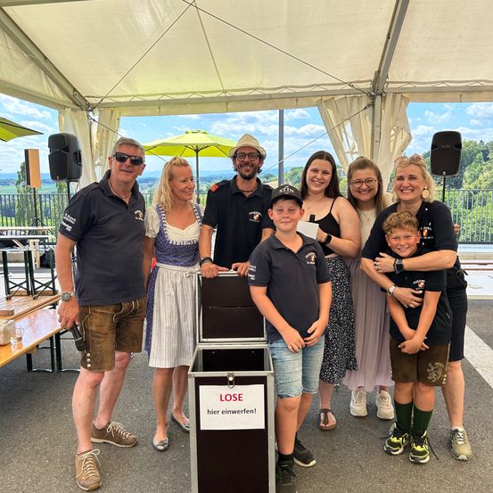 Eine Gruppe von Menschen posiert für ein Foto unter einem Zelt, einer hält eine Spendenbox mit der Aufschrift 'LOSE hier einwerfen!' (werfen Sie Ihr Kleingeld hier rein). Sie tragen lässige Sommerkleidung. Hinter ihnen steht ein Holztisch mit einem Glas, und zwei Lautsprecher sind sichtbar.