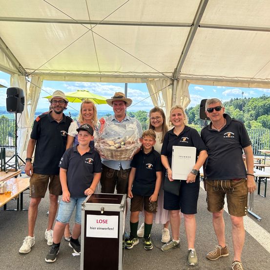 Eine Gruppe von Menschen, einschließlich Erwachsener und Kindern, steht unter einem Zelt mit einem Korb Brot. Eine Frau hält eine Urkunde, und ein Mann steht neben einer Box mit der Aufschrift 'LOSE'. Im Hintergrund befinden sich Berge und ein Zaun.