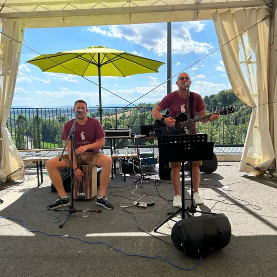 Zwei Männer spielen auf einer Terrasse mit einem gelben Regenschirm, einer spielt Gitarre und singt, der andere sitzt und singt. Hinter ihnen bietet ein Metallzaun einen Blick auf eine hügelige Landschaft.