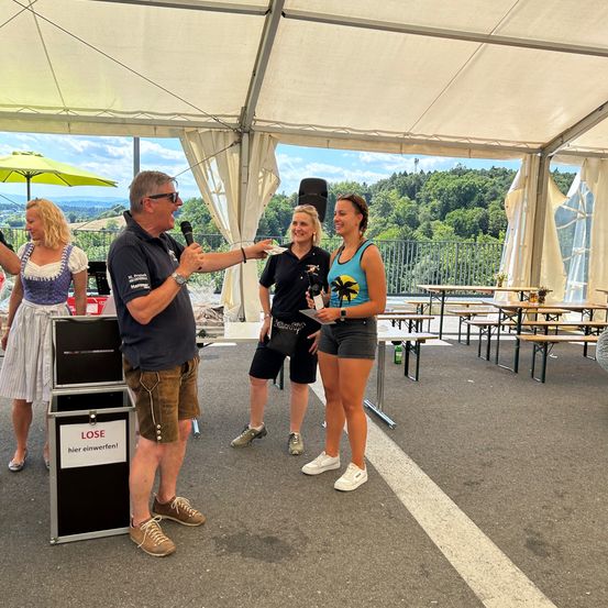 Ein Mann spricht in ein Mikrofon und steht unter einem Zelt mit zwei Frauen. Sie befinden sich in einem Park mit Tischen und Bänken. Bäume und Berge sind im Hintergrund.