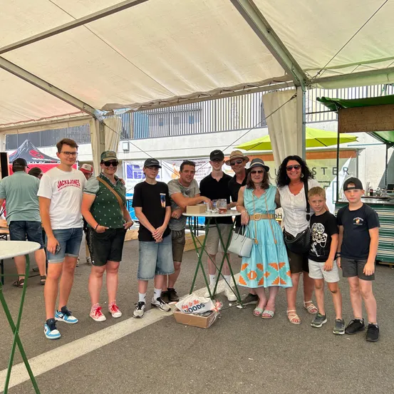 Eine Gruppe von Menschen posiert für ein Foto unter einem Zelt, mit einem Tisch davor. Sie tragen alle Freizeitkleidung. Der Tisch hat einige Getränke und Essensartikel. Auf dem Boden befinden sich einige Lagerboxen. Dahinter steht ein Zelt mit einem grünen Dach.