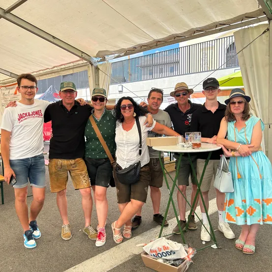 Eine Gruppe von Menschen, darunter zwei Frauen, posiert für ein Foto unter einem Zelt. Sie tragen Freizeitkleidung. Es gibt einen Tisch mit einem Glas und einer Schachtel. Hinter ihnen steht ein Metallzaun.