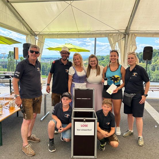 Eine Gruppe von Menschen posiert für ein Foto unter einem Zelt, wobei eine Person kniet und eine andere eine Flasche hält. Dahinter steht ein Tisch mit Getränken und eine Aufbewahrungsbox mit der Aufschrift 'LOSE hier einwerfen!'.