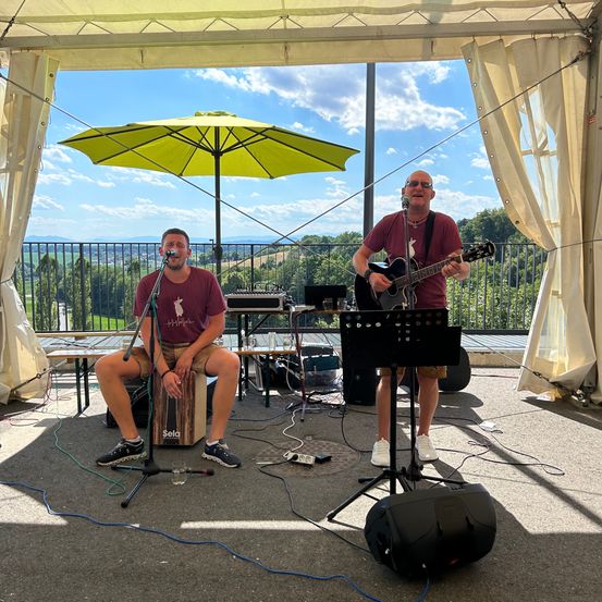 Zwei Männer spielen auf einer Freiluftbühne, einer sitzt mit einem Mikrofon und der andere spielt Gitarre. Hinter ihnen steht ein gelber Schirm, Lautsprecher und eine malerische Aussicht.
