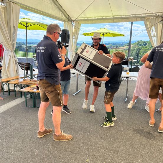 Ein Mann mit Mikrofon hilft einem Jungen, eine Kiste unter einem Zelt zu heben, während andere stehen und Berge im Hintergrund sind.
